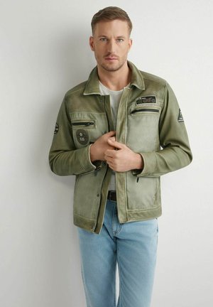 Mężczyzna zapinający zieloną, wojskowego stylu kurtkę na białą koszulkę, ubrany w jasnoniebieskie jeansy, stojący na jednolitym jasnym tle.