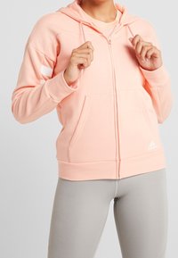Felpa con zip di un delicato colore rosa chiaro, realizzata in tessuto morbido, con tasca a marsupio, cordini sul cappuccio e accento del logo Adidas bianco.