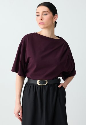 Donna che indossa una maglia larga di colore viola scuro con maniche lunghe fino al gomito, una gonna nera e una cintura nera con fibbia ovale dorata.