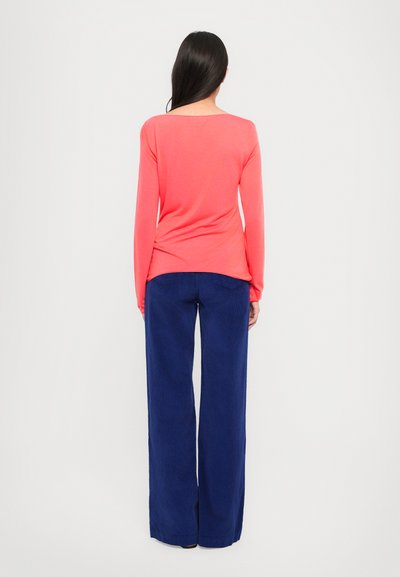 Maglietta corallo a maniche lunghe con scollo ampio, abbinata a pantaloni blu navy a vestibilità morbida realizzati in tessuto texturizzato, vista da dietro.