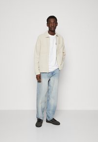 Jachetă din fleece bej deschis, cu fermoar, cu două buzunare frontale, purtată peste un tricou alb, asortată cu blugi lejeri de denim albastru deschis și adidași negri.
