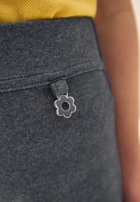 Hallid puuvillased püksid, millel on sile tekstuur ja vöökohal dekoratiivne hõbedane lillekujuline luku tõmbaja. Kollane riie on ülal nähtav.