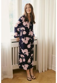 Duga mornarsko plava haljina s velikim ružičastim cvjetnim uzorkom, kopčanjem na drukere i zvonastim rukavima, uparena s crnim japankama na drvenoj podlozi.