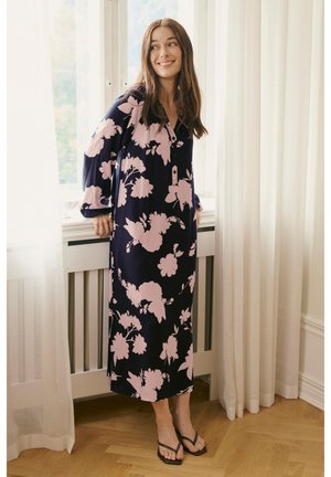 Lang navyblå kjole med stort pink blomsterprint, knaplukning og klokkeærmer, kombineret med sorte flip-flops på et trægulv.