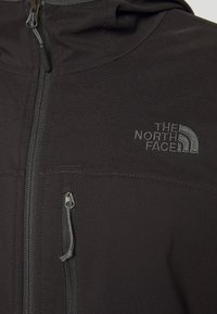 Veste noire à fermeture éclair avec poche zippée sur le devant et logo brodé "The North Face" sur le côté droit de la poitrine.