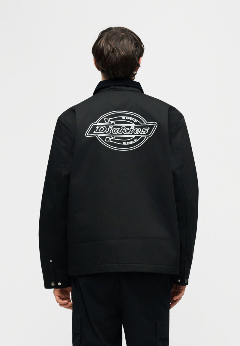 Persona vista da dietro che indossa una giacca nera con un grande logo bianco "Dickies" sul retro, su uno sfondo chiaro e uniforme.