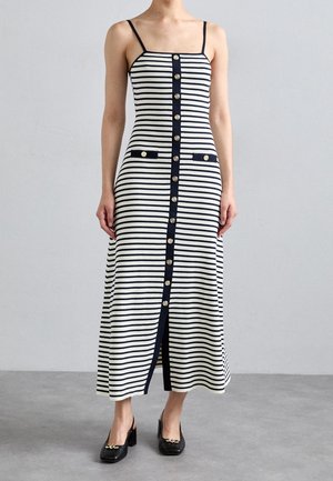 Robe midi sans manches pour femme, rayée horizontalement noir et blanc, avec des boutons dorés au centre et des chaussures noires à talons blocs.