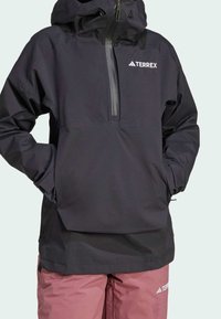 Veste TERREX noire avec une fermeture éclair sur le devant et une capuche, en tissu lisse et imperméable, dotée de poignets élastiques. Associée à un pantalon rose clair.