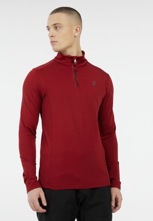 Junger Mann in einem roten Langarm-Quarter-Zip-Pullover und schwarzen Hosen, der nach links schaut, vor einem weißen Hintergrund.
