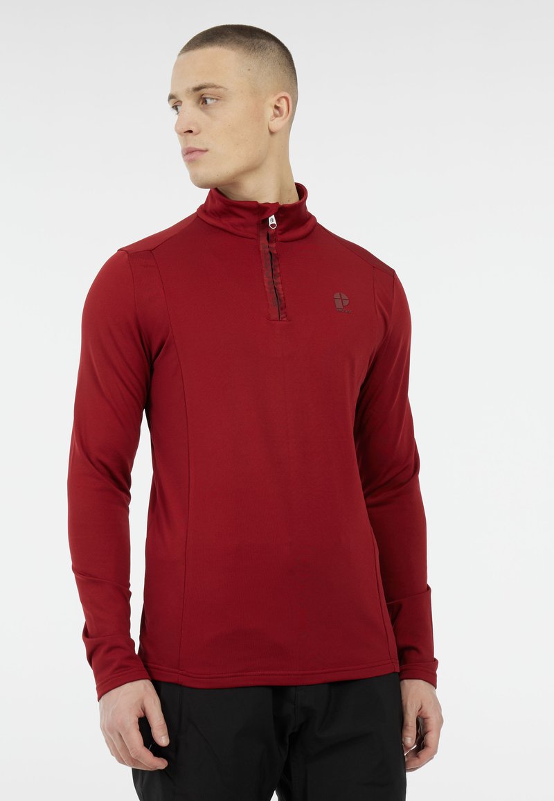 Giovane uomo che indossa un pullover rosso a maniche lunghe con zip a un quarto e pantaloni neri, guardando verso sinistra su uno sfondo bianco.