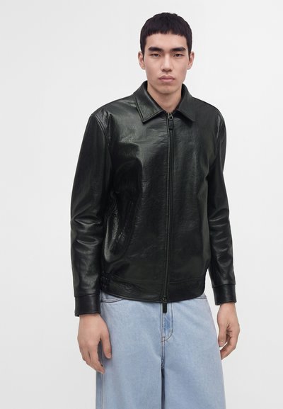 Young man wearing a black leather zip-up jacket and light blue jeans, standing against a plain white background.