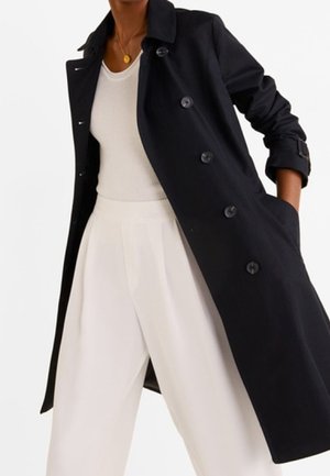 Osoba ubrana w czarny dwurzędowy płaszcz przeciwdeszczowy na białym, bezrękawnym topie i białych, szerokich spodniach z wysokim stanem.