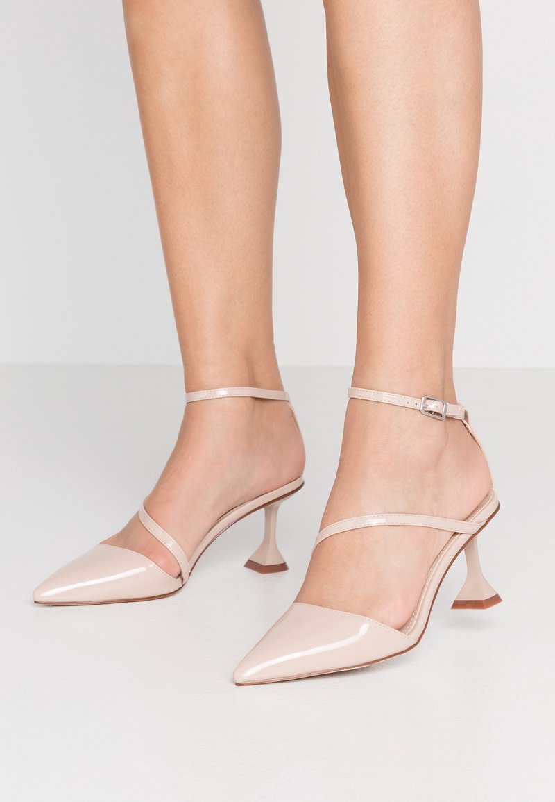 Jambes nues portant des escarpins en cuir verni rose clair à bout pointu avec des fines brides à la cheville et à l’avant sur un fond uni.