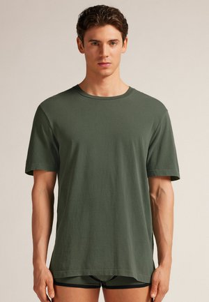 Camiseta de algodón verde oscuro con cuello redondo y mangas cortas. Textura suave, corte holgado y dobladillo recto. Llevada sobre pantalones cortos a juego.