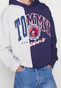 Kleurvlakken hoodie in lichtgrijs en marineblauw, met grote "TOMMY" logo's. De stof is zacht, met een ontspannen pasvorm en een buidelzak.