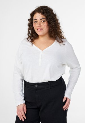 Donna con i capelli ricci e lentiggini che indossa una camicia henley bianca a maniche lunghe e pantaloni neri a vita alta, in piedi davanti a uno sfondo bianco.