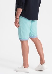 Pantaloni corti in cotone azzurro chiaro con risvolti, abbinati a sneakers bianche. La vista posteriore mostra una vestibilità rilassata e un design pulito.