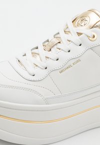 Baskets compensées blanches avec accents dorés, lacets blancs et marquage "Michael Kors" sur le côté et la languette.