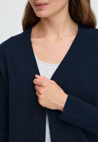 Cardigan bleu marine en maille, avec un décolleté en V profond, des manches longues et une coupe décontractée. Vue rapprochée du col et de la main.