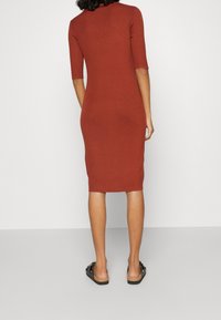 Robe en maille côtelée terracotta, longueur mi-mollet, avec manches courtes et silhouette ajustée. Portée avec des sandales slides noires.