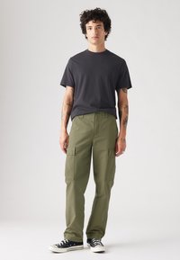 T-shirt cinza escuro, com corte cropped, combinado com calças cargo verde-azeitona com grandes bolsos laterais e sapatilhas pretas de cano baixo com sola branca.