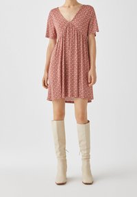 Rotes, kurzes Blumenkleid mit kurzen Ärmeln, V-Ausschnitt, gerafftem Oberteil und fließendem Schnitt. Kombiniert mit knielangen, beigen Stiefeln.