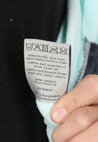 Label for fleece garment in gray fabric, showing care instructions and symbols. Light teal fleece edge visible. Hand holding the label.