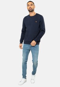Marine crewneck sweatshirt met oranje logo-detail, gecombineerd met lichtblauwe skinny jeans en witte sneakers. Katoenen materiaal, casual ontwerp.