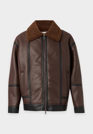 Homme portant une veste en cuir marron avec col en peau lainée, chemise couleur rouille, jean beige large et chaussures beiges sur fond blanc uni.