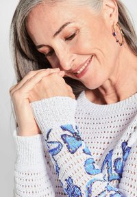 Weißer Strickpullover mit blauen und schwarzen Blumenstickereien an den Ärmeln. Die Textur ist offen gestrickt und verfügt über eine lockere Passform.