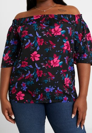 Kvinde iført sort off-shoulder bluse med pink, rød og lilla blomsterprint, kombineret med blå jeans og en diamant-tatovering på skulderen.