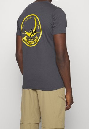 Mężczyzna w ciemnoszarej koszulce z dużym żółtym logo drużyny wspinaczkowej i sylwetką na plecach, ubrany w beżowe szorty cargo.