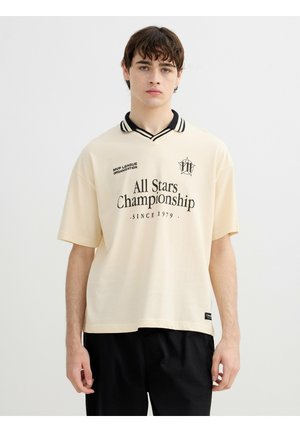 Joven con camiseta polo beige con cuello negro, texto negro "All Stars Championship Since 1979" y pantalones negros, de pie sobre fondo blanco.