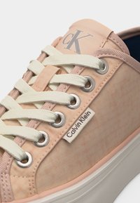 Κοντινή λήψη ενός παπουτσιού Calvin Klein σε ροδακινί απόχρωση με λευκά κορδόνια, ασημένιες οπές, και εμφανή λογότυπα "CK" και "Calvin Klein" στο ύφασμα.