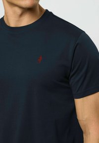 Vyras dėvi tamsiai mėlynį trumpomis rankovėmis marškinėlį su mažu raudonu siuvinėtu figūra kairėje krūtinės pusėje.