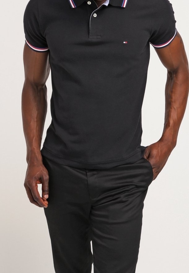 Homme portant un polo noir avec des bordures rouges, blanches et bleues, et un pantalon noir, une main dans la poche.