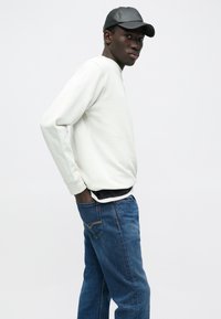 Világosszürke pulóver bordázott mandzsettákkal és szegéllyel, fekete ing fölött viselve, kék, egyenes szárú farmerrel párosítva. Fekete, fényes sapka.