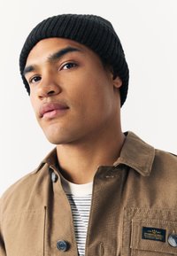 Smeđa platnena jakna s crnom rebrastom pletenom kapom, slojevito preko prugaste košulje. Ima vidljive gumbe i oznaku na džepu.