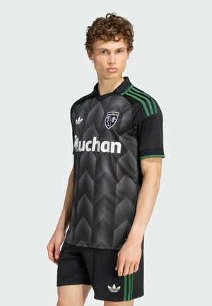 Joven con cabello rizado que lleva un uniforme de fútbol negro y verde de Adidas con patrones geométricos y el logo de RCL, posando frente a un fondo liso.