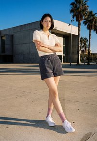 Jeune femme debout, bras croisés, portant une chemise blanche, un short foncé, des chaussettes roses et des baskets blanches sur un trottoir ensoleillé avec des palmiers.