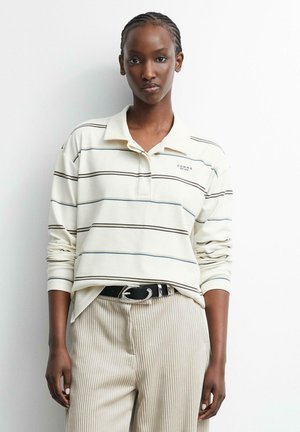 Junge Frau mit geflochtenem Haar trägt ein cremefarbenes, langärmeliges Poloshirt mit dünnen horizontalen Streifen und beige gerippte Hosen mit schwarzem Gürtel.