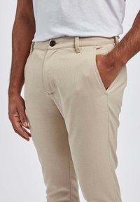 Mężczyzna ubrany w beżowe spodnie i białą koszulę, z jedną ręką włożoną do kieszeni spodni, a drugą swobodnie opuszczoną wzdłuż ciała.