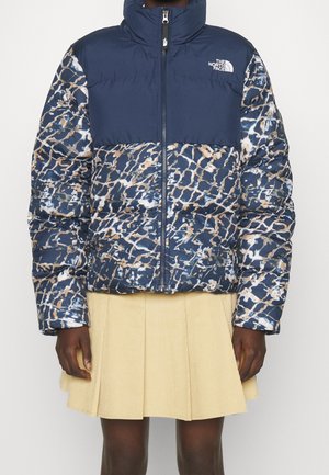 Marineblauw en een patroon: de pufferjas van The North Face met abstracte print, gedragen met een beige geplooide rok op een donkerhuidige persoon tegen een effen achtergrond.