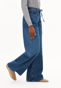 Jean bleu à jambe large en denim, doté d'une taille haute, d'un nœud à l'avant et de détails légèrement déchirés. Associé à des chaussures de couleur claire.