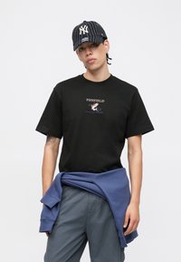 Black cotton t-shirt with a printed logo in the center. Model wears a dark blue hoodie tied around the waist and a black striped cap.