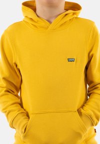Sudadera de felpa amarilla con bolsillo tipo canguro, puños acanalados y un pequeño parche de logo verde de Levi's en el pecho.