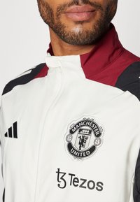 Witte sportjack met een kastanjebruine kraag en zwarte accenten. Beschikt over geborduurd Manchester United-logo en Tezos-merkbewerking.