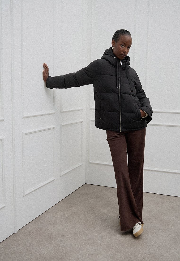 Chaqueta de plumas negra con cremallera frontal y capucha con cordón, combinada con pantalones acampanados marrones y zapatillas de color claro, sobre un fondo neutro.