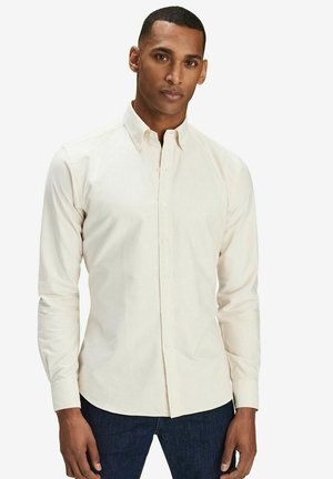 Ljusbeige skjorta med knappar och långa ärmar, tillverkad av bomull, med spetsig krage och en figurnära design. Bärs tillsammans med mörk denimjeans.