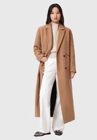 Καμηλό παλτό από μαλλί με κανονικό γιακά και διπλό μπροστινό. Συνδυασμένο με κρεμ πουλόβερ με πλέξη και άσπρα παντελόνια με φαρδιά γραμμή.